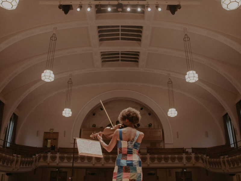 Almaz Ohene playing the violin in a large building with arched ceilings and large windows on either side